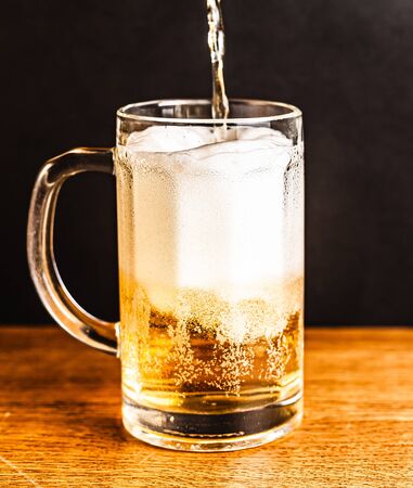 Cold beer with foam in a mug, on a wooden table and a dark background with blank space for  text. Stock Photo mug of cold foamy beer close-up.の写真素材