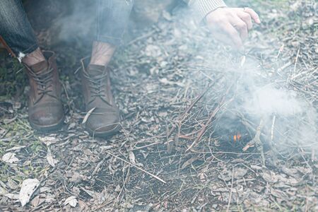 A stock photo of a man in nature sitting by a small fire, warming himself.