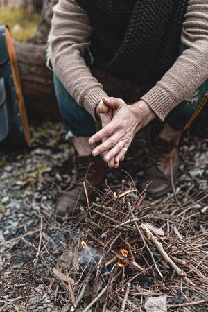 A stock photo of a man in nature sitting by a small fire, warming himself.