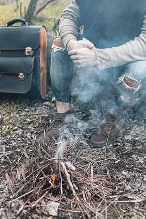 A stock photo of a man in nature sitting by a small fire, warming himself.
