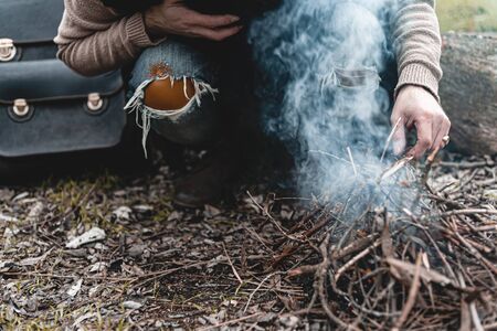A stock photo of a man in nature sitting by a small fire, warming himself.