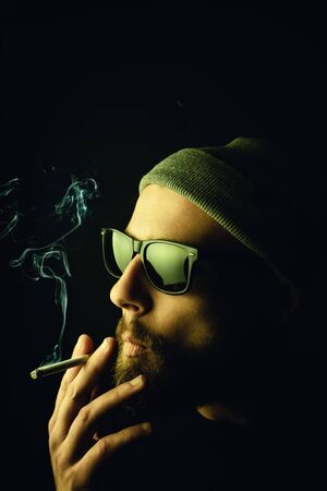 Portrait of a bearded, white hipster smoker on a dark background. Studio stock photo concept of smoking cigarettes and marijuana.の写真素材