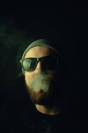 Portrait of a bearded, white hipster smoker on a dark background. Studio stock photo concept of...