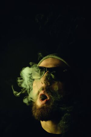 Portrait of a bearded, white hipster smoker on a dark background. Studio stock photo concept of smoking cigarettes and marijuana.の写真素材