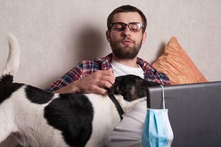 The guy works at home on freelance with his pet. The guy interacts with a dog on the couch with a medical mask.の写真素材