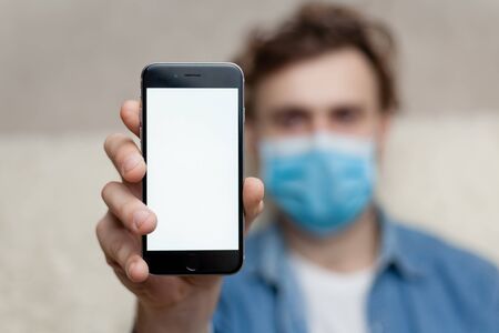 The guy shows a blank smartphone display in a medical mask at home. Concept coronavirus quarantine and application for phone or call medical service.の写真素材