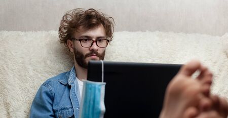 Handsome guy working on a laptop while lying on a sofa. Remote work and study at home. The concept of quarantine and social distance during quarantine.の写真素材