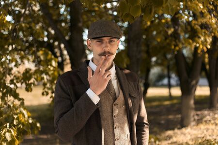 Retro 1920s portrait of an English gangster with a flat cap. Smokes a cigarette on the street..