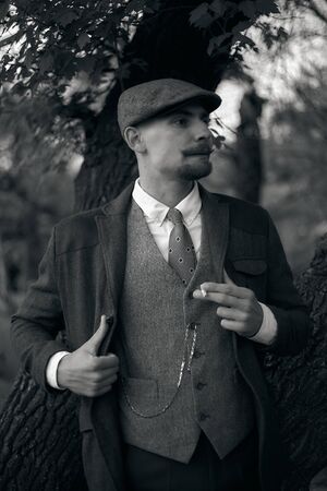 Retro 1920s portrait of an English gangster with a flat cap. Smokes a cigarette on the street..