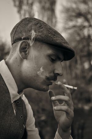 Retro 1920s portrait of an English gangster with a flat cap. Smokes a cigarette on the street..の写真素材