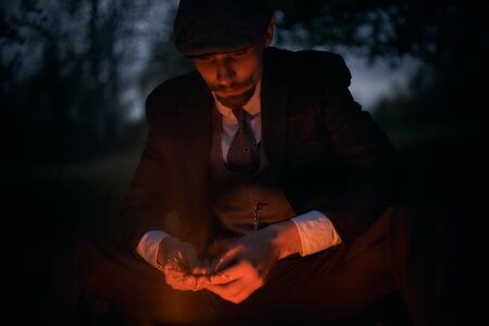 Portrait of a guy sitting by the fire at night in nature. Cinematic vintage photo. Retro 1920s...