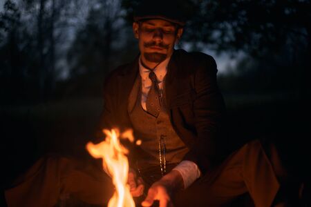 Portrait of a guy sitting by the fire at night in nature. Cinematic vintage photo. Retro 1920s...