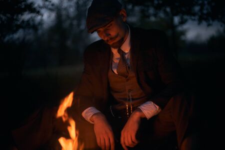 Portrait of a guy sitting by the fire at night in nature. Cinematic vintage photo. Retro 1920s portrait of an English man with a flat cap.の写真素材