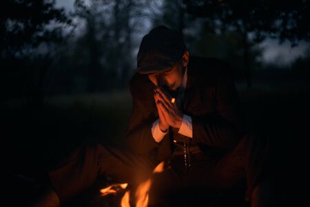 Portrait of a guy sitting by the fire at night in nature. Cinematic vintage photo. Retro 1920s...