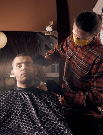 Male hairdresser in a protective mask and gloves, dressed in casual clothes. Client without a mask. Hairdressing after quarantine. Health precautions. Small business. Coronavirus pandemic aftermath.の写真素材