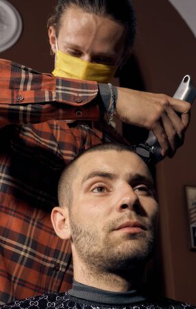 Male hairdresser in a protective mask and gloves, dressed in casual clothes. Client without a mask. Hairdressing after quarantine. Health precautions. Small business. Coronavirus pandemic aftermath.の写真素材