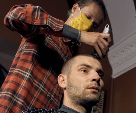Male hairdresser in a protective mask and gloves, dressed in casual clothes. Client without a mask. Hairdressing after quarantine. Health precautions. Small business. Coronavirus pandemic aftermath.の写真素材