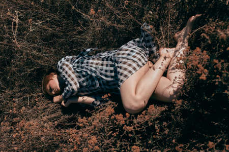 Attractive girl resting in the yellow dry grass in a dress barefoot. Model in a plaid dress.