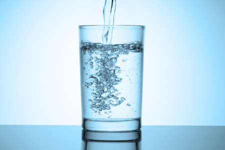 Jet of water fills a glass stands on a glossy table with reflection. Water splash with bubbles. Cold blue tinted vignette.の写真素材