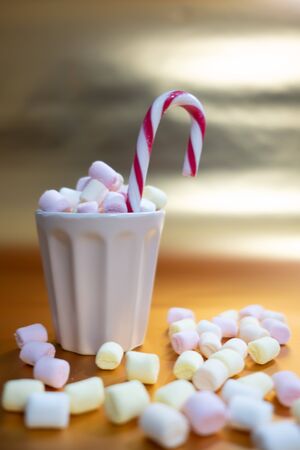 Christmas background with a cup full of marshmallows and a candy cane, marshmallows on the table surface and bright golden backgroundの写真素材