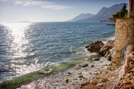 A rocky seashore with a stone building and mountains in the backgroundの写真素材