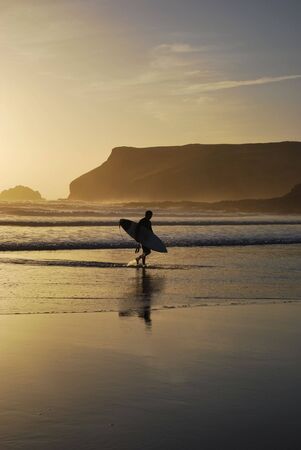 A surfer leaves the surf as the sun sets at Polzeath Beach in Cornwall, UK.の写真素材