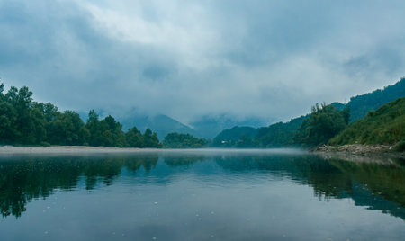A cloudy morning on the Drina River. Mountains hiding in low clouds and a pair of fishermen on the bank of a fog-covered riverの写真素材