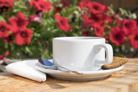 Cup of coffee served on a wooden wood table with a white napkin, spoon and biscuit with a blurred floral backgroundの写真素材