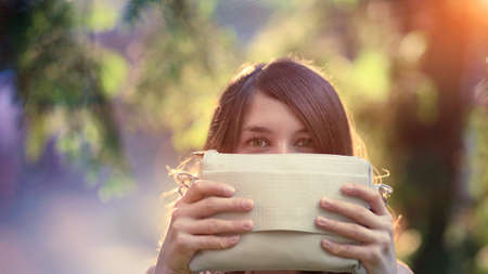 Funny teenage girl hidding by holding a beige purse in front of her face, outdoors in a sunny day of summerの写真素材