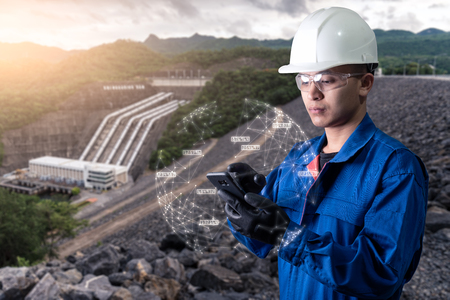 The abstract image of the engineer holding smartphone with hologram and the hydroelectric dam is backdrop. the concept of clean energy, hydroelectricity, water management and engineering.の写真素材