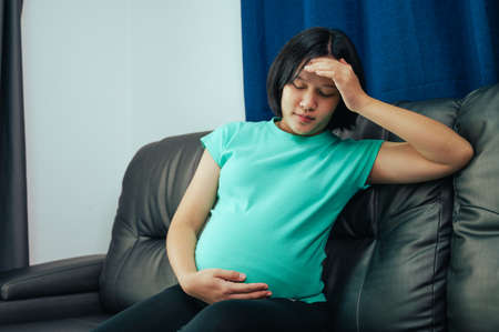 Asian pregnant woman suffering from headache while sitting on sofa at home.の写真素材