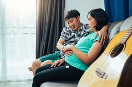 Happy asian pregnant couple sitting on sofa with guitar at home.の写真素材