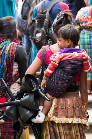 SAN JUAN OSTUNCALCO, GUATEMALA - JUNE 24: An unidentified woman vendor in traditional dress carrying a child in a sling on her back and selling goods through crowds at the San Juan Ostuncalco fair in honor of Saint John the Baptist on June 24, 2017 in Guaのeditorial素材