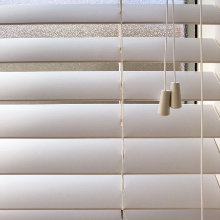 White wooden window blinds partially open with adjustment cords inside a houseの写真素材