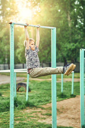 Full body side view of focused preteen boy in casual clothes doing exercises on metal bar while spending summer day on sports ground in parkの写真素材