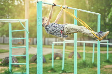 Full body side view of focused preteen boy in casual clothes doing front level with a fitness elastic band on metal bar while spending summer day on sports ground in parkの写真素材
