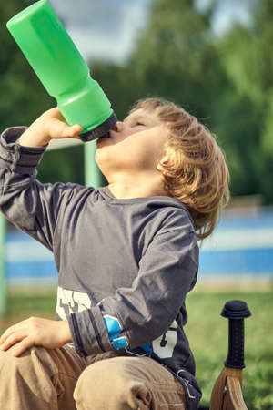 Tired little boy in casual clothes with wooden balance bike drinking water from plastic bottle while sitting on green grass of playground in summer dayの写真素材