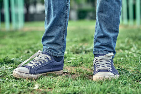 Crop anonymous male child in jeans and gumshoes standing on green grass on playground while spending...