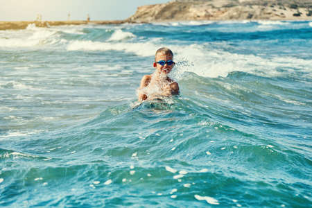 Shirtless boy in swimming goggles standing in sea water and playing with foamy waves on sunny summer...