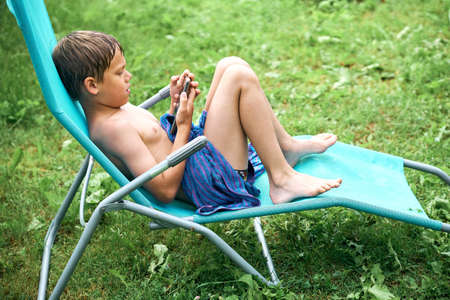 Gadget addiction. Wet boy sitting on sun lounger and playing on smartphone after swimming in outdoor poolの写真素材