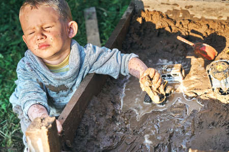 Cute serious preschooler boy with dirty face and clothes playing with toy cars in the sandbox near wooden rural house in summer day in countrysideの写真素材