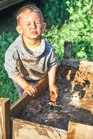 Cute serious preschooler boy with dirty face and clothes playing with toy cars in the sandbox near...
