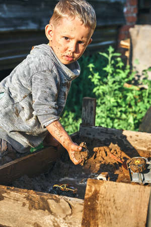 Cute serious preschooler boy with dirty face and clothes playing with toy cars in the sandbox near...
