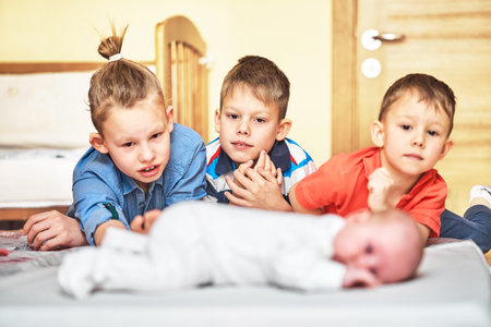 Funny brothers wearing bright clothes look seriously at defocused sweet baby in white body lying on fluffy sheet in foregroundの写真素材