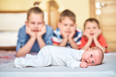 Blurry brothers in bright clothes look seriously at sweet sibling in white body lying on baby changing table sheet closeupの写真素材