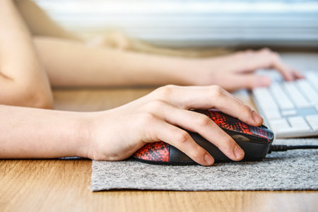 Young schoolboy hands on red black mouse with felt pad and blurred keyboard of white color on brown...