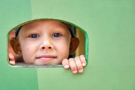 Cute happy little boy looks through small window in green wooden house spending time on playground on summer day close viewの写真素材