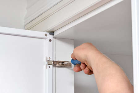 Skilled handyman adjusts furniture hinge with screwdriver installing contemporary white case in...