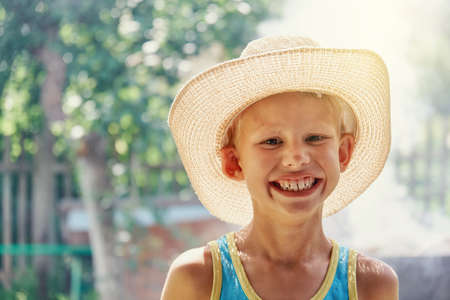 Cheerful little boy wearing large straw hat and blue sleeveless shirt posing for camera and smiles all over his mouth in sunny summer city park close viewの写真素材