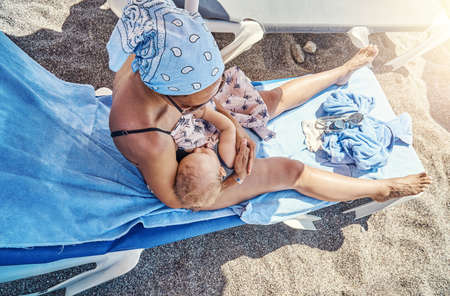 Young woman with blue bandana on head holds cute sleeping baby in arms sitting on deckchair with towel on sand beach upper viewの写真素材